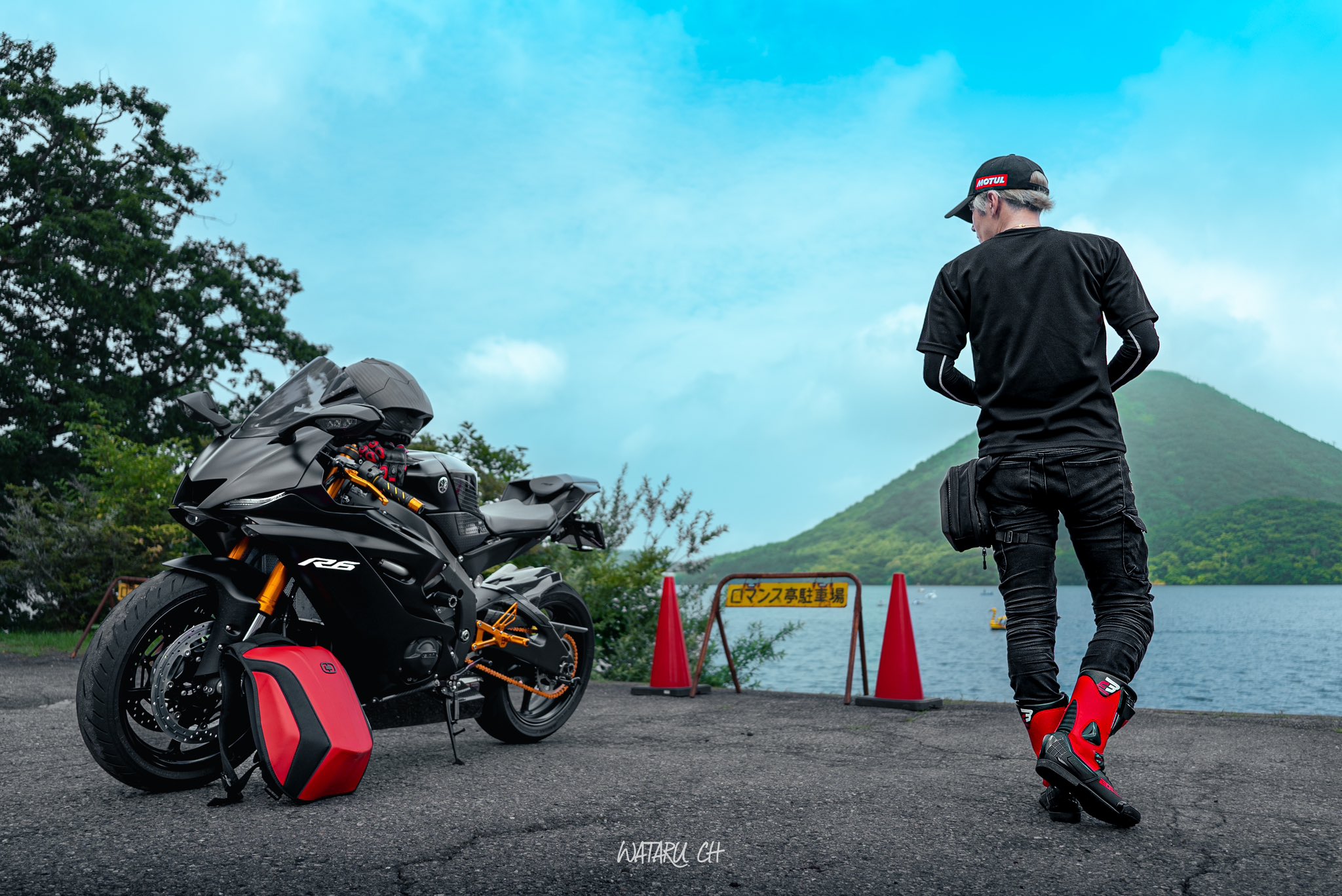 a man in black clothing and red boots stands near his black yamaha r6 motorcycle with a red helmet. he is standing on a paved road with a sign behind him. he is looking out at a lake with a mountain in the background. the sky is blue with white clouds.