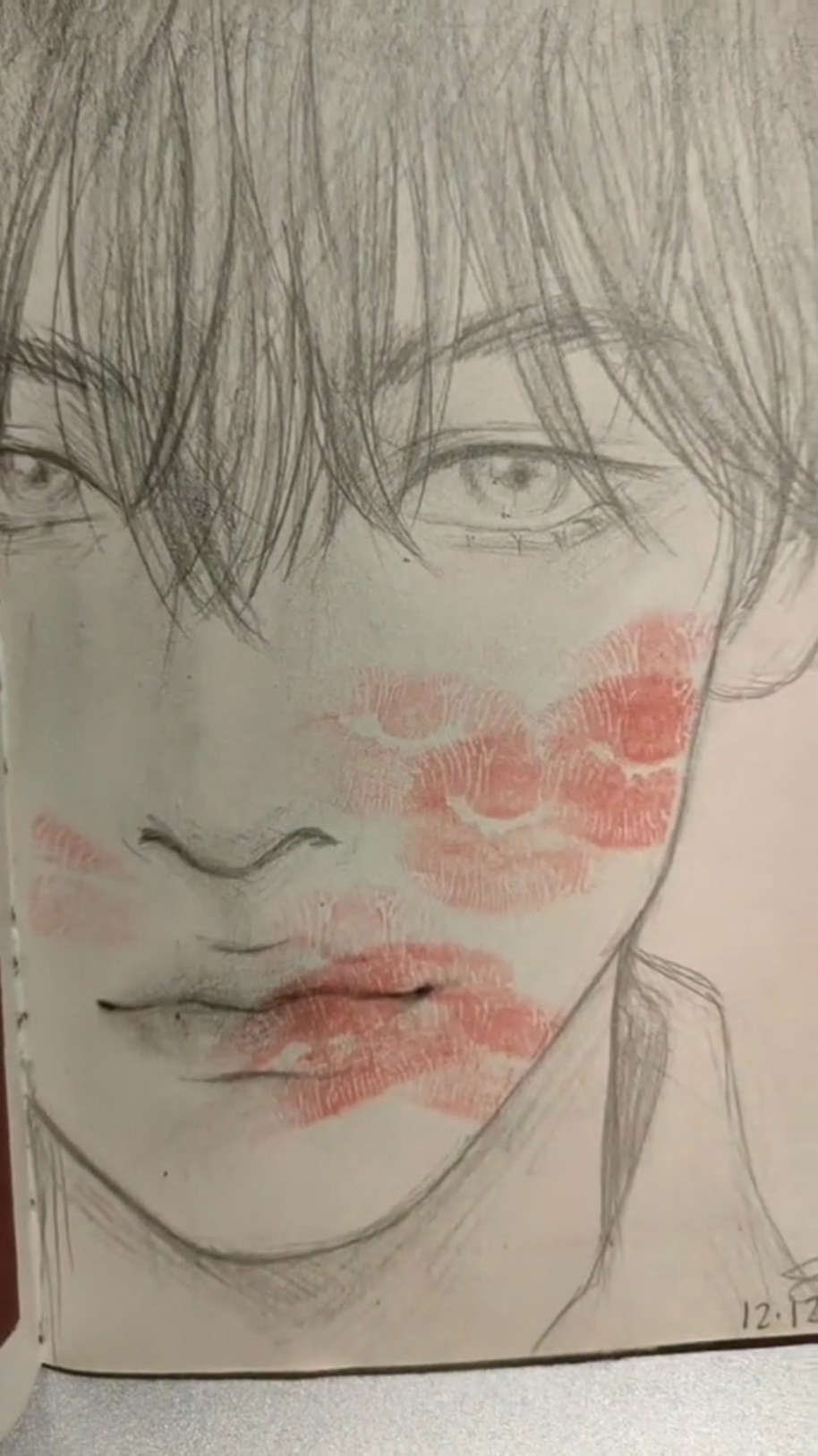 a realistic pencil sketch of a man's face with lipstick kisses in pink and red on his cheeks, jawline, and mouth.  the drawing appears to be in a sketchbook, and the lighting is soft.