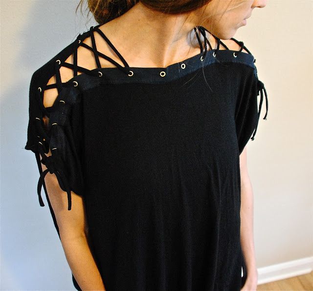 a woman is wearing a black laceup tshirt. the tshirt has eyelets along the shoulders with laces threaded through them. the sleeves also feature a laceup design. the woman is standing against a lightcolored wall.