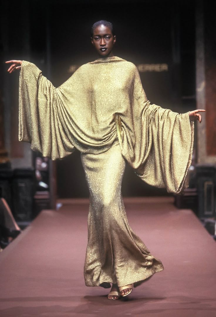 a black woman wearing a gold, floorlength dress with cape sleeves is walking down a runway. she has her arms outstretched and is wearing sandals. the background is blurry, but it appears to be a fashion show setting.