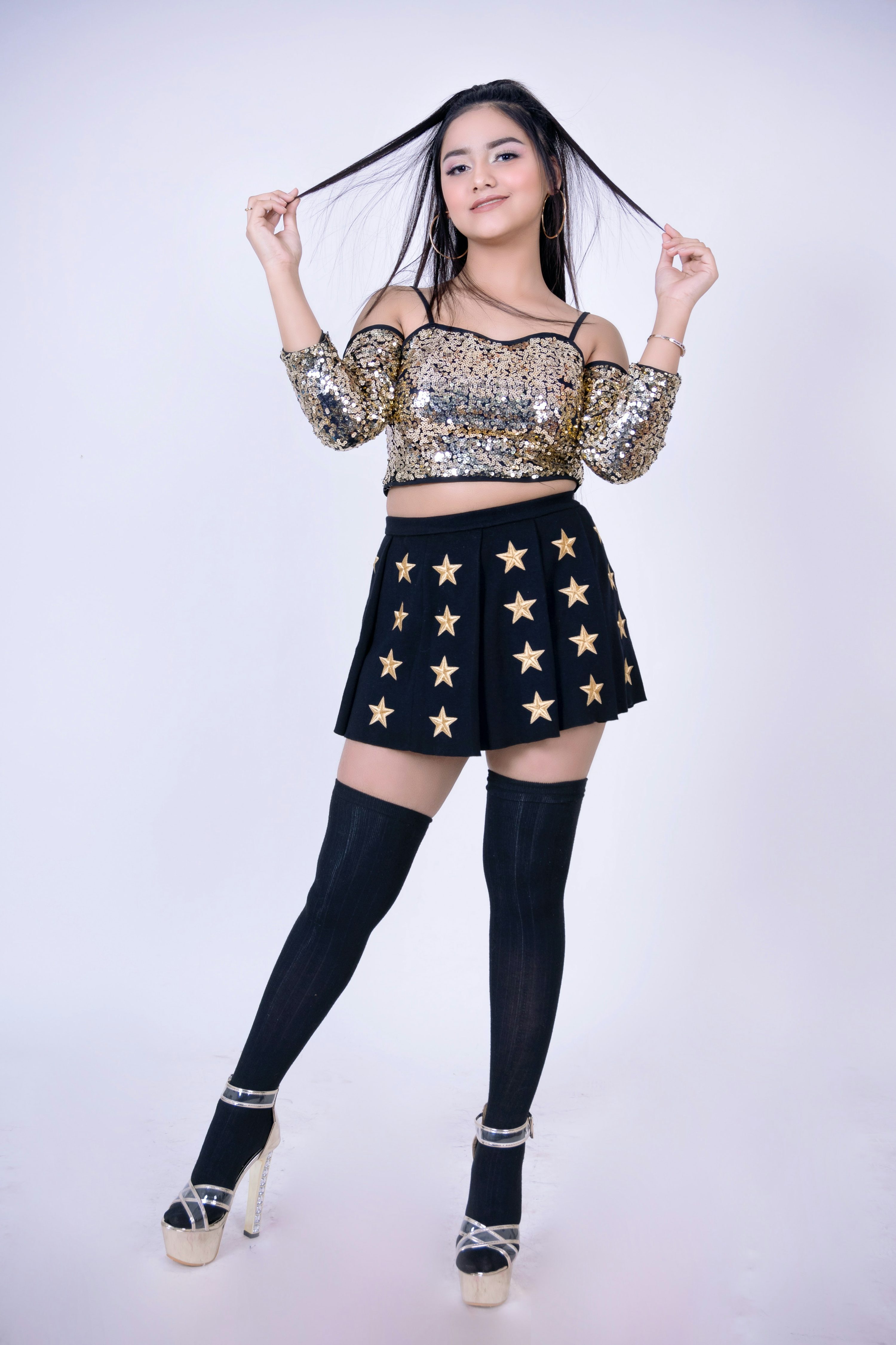 a young asian woman poses confidently in a studio against a white background. she is wearing a gold sequin crop top, a black mini skirt with gold star embellishments, black kneehigh socks, and highheeled sandals. her long black hair is styled loosely, and she has on makeup. she looks directly at the camera with a happy and confident expression.  the overall style is trendy and stylish.