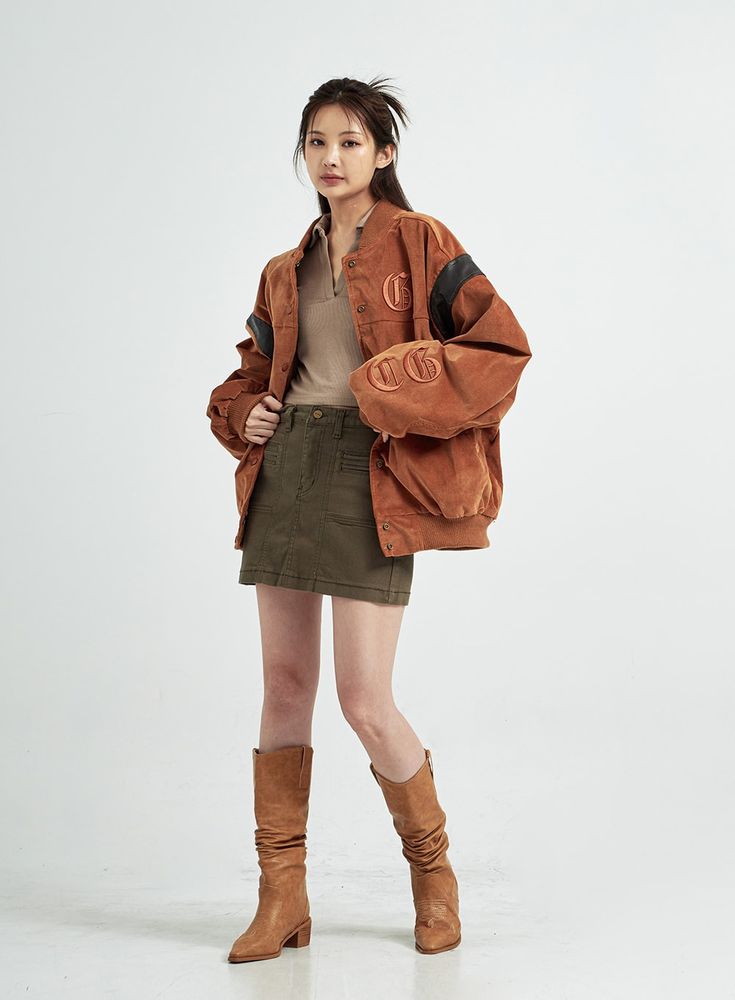 a fullbody shot of a young woman wearing a brown corduroy bomber jacket with black accents and embroidered details, a khaki mini skirt, and tan slouch boots. she's posing against a light gray background.  her hair is styled in a sleek updo. the overall style is casual yet fashionable, suitable for autumn or fall.