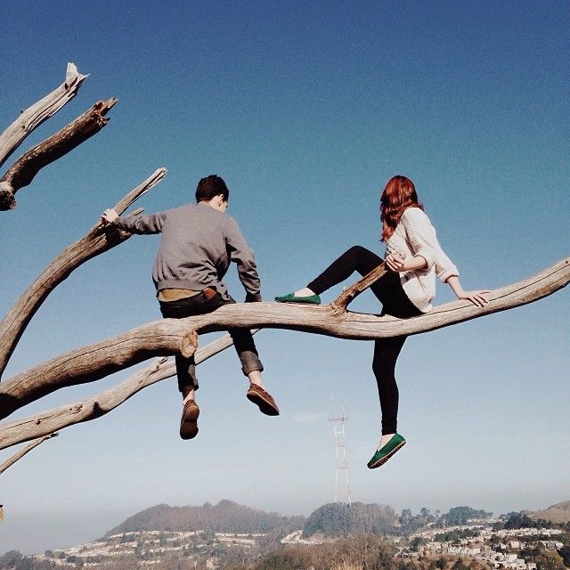 a couple sitting on a tree branch, looking out at the city. the woman is wearing a red shirt and has long red hair. the man is wearing a gray shirt. they are both smiling and seem to be enjoying the moment together. the background is a city skyline with a mountain range in the distance. it is a sunny day, and the sky is blue. the photo is taken from a low angle, giving a sense of perspective. it is a  romantic image that captures the beauty of a couple's love and the joy of being together.