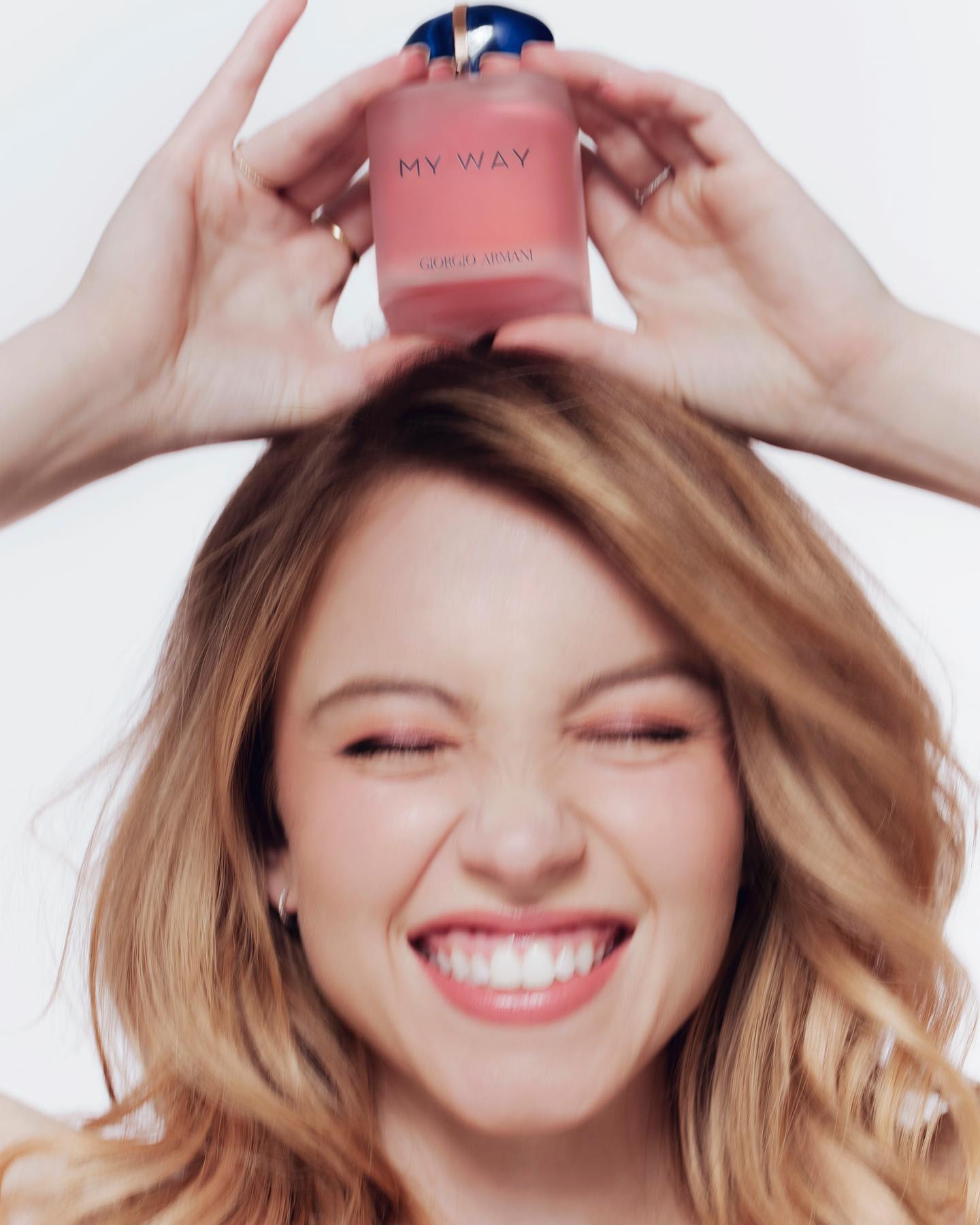 a closeup shot features a smiling blonde woman holding a giorgio armani my way perfume bottle atop her head. the woman is the focal point, with her happy expression and wavy hair. the perfume bottle is prominently displayed, showcasing its pink hue and branding.  the background is a simple, uncluttered white, emphasizing the model and product. the overall aesthetic is one of beauty, elegance, and luxury, consistent with highend perfume advertising.