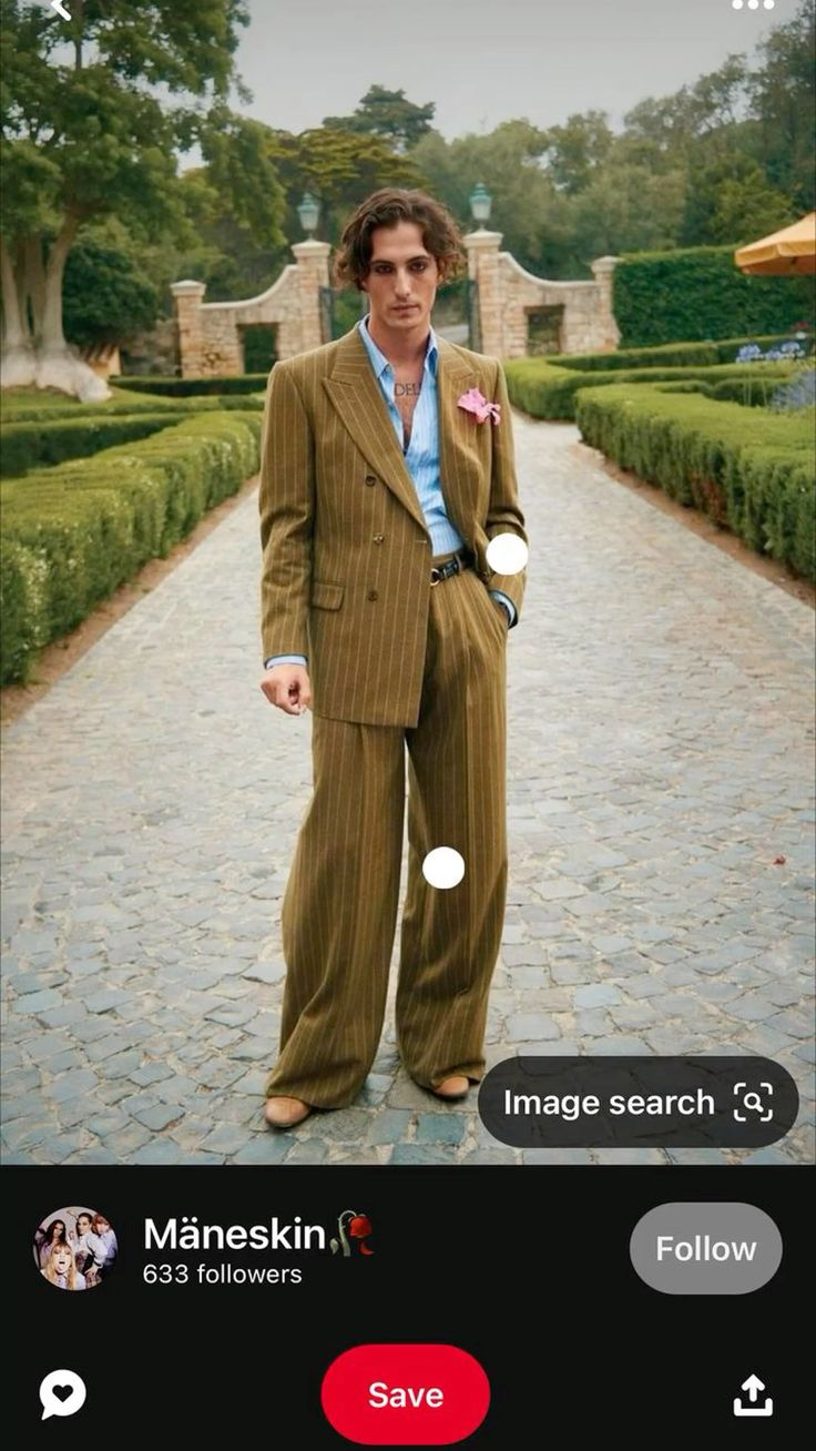 a man in a pinstripe suit is standing on a stone path, looking at the camera. the man is wearing a blue shirt and a dark tie, with a pink flower on his suit lapel. his shoes are brown leather. the background is a lush garden with green plants and trees. the man is standing in the middle of a walkway lined with cobblestones.