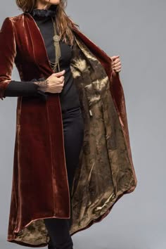 a woman wearing a long, red velvet coat with gold lining. she is standing with her arms crossed and looking slightly to the side. the coat is flowing and elegant, and it looks very comfortable. the woman's hair is pulled back in a simple style, and she is wearing a black top and pants underneath the coat. the background is a plain, white wall, so the focus is on the coat.