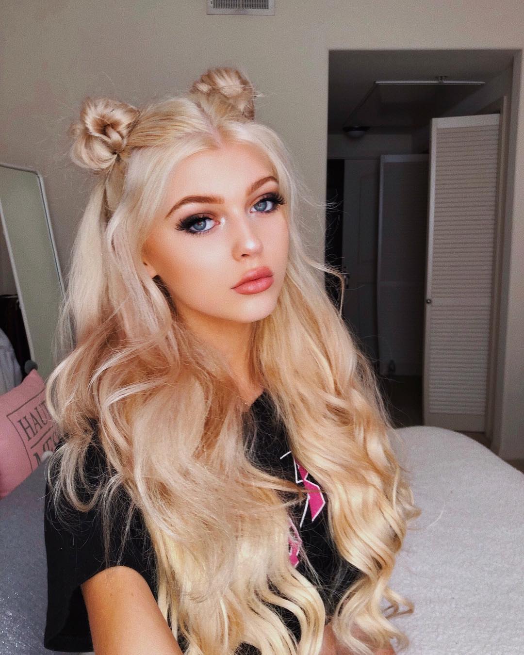 a young woman with long, wavy blonde hair styled in two buns on top of her head poses for a portrait. she has blue eyes and is wearing makeup. she is in a bedroom setting with a bed behind her.