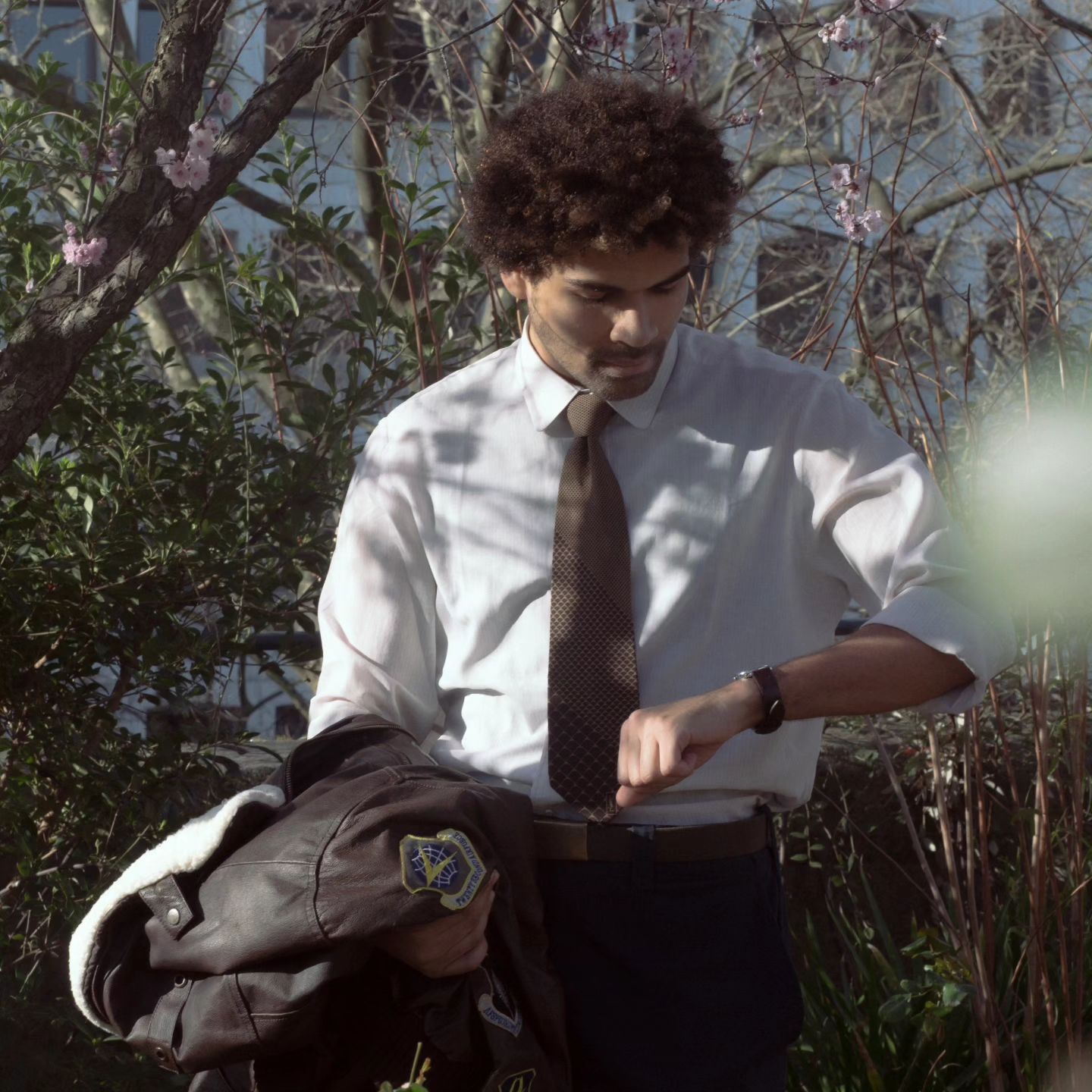 a young man with an afro hairstyle, wearing a white shirt, tie, and dark pants, is standing outdoors in a garden setting. he is checking the time on his wristwatch while holding a brown leather bomber jacket with a patch on it. the image suggests he might be running late for something. the lighting is natural and overcast, with the focus on the man and his surroundings.
