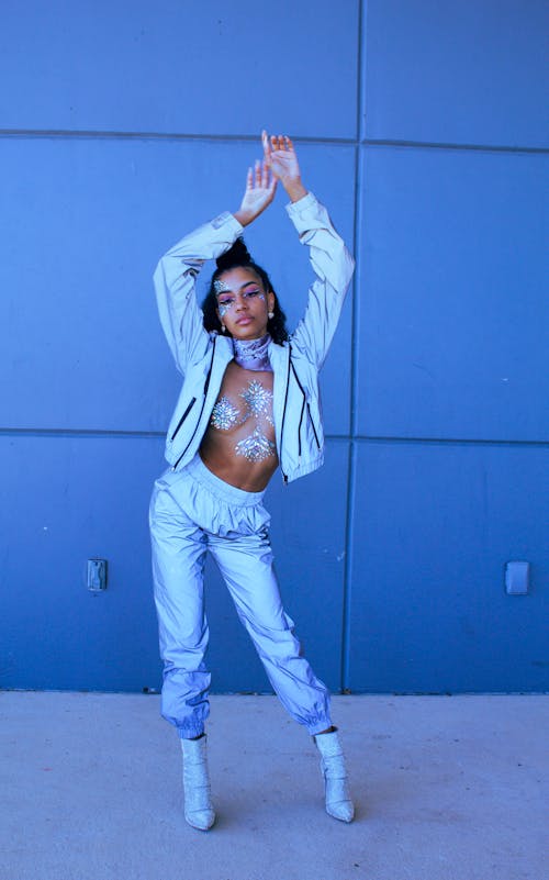 a young woman in a stylish silver tracksuit stands against a blue wall, arms raised.  she is wearing a silver jacket and matching pants, along with silver boots. she has glitter body paint on her chest and subtle makeup.  the overall style is contemporary and trendy. the image is welllit and the woman appears confident.