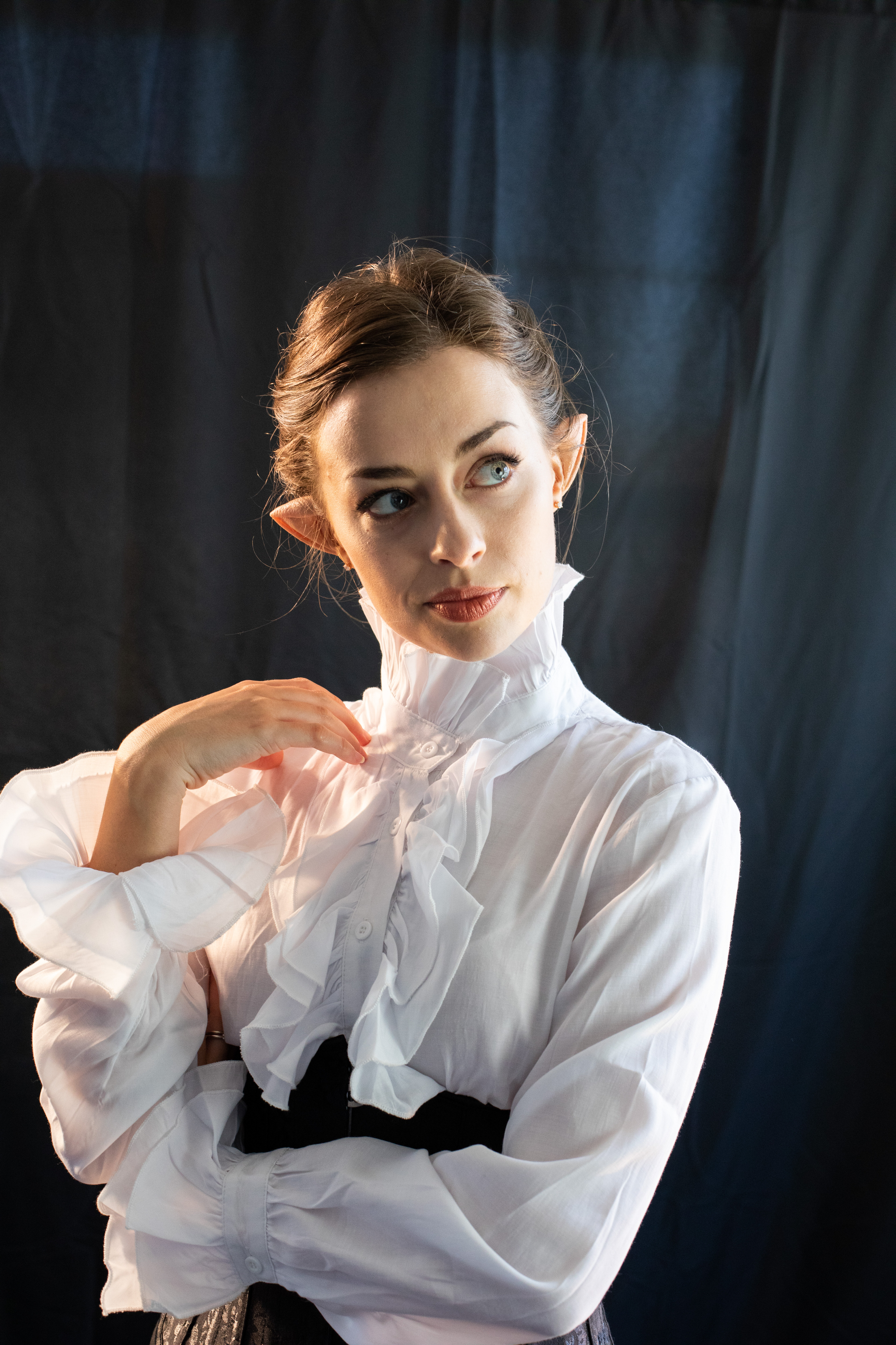 closeup portrait of a woman with elf ears, dressed in an elegant white ruffled blouse with a high victorianstyle collar. she's posing against a dark backdrop, arms crossed, and her expression is thoughtful and slightly pensive as she looks off to the side. the image has a dramatic and theatrical feel, possibly suggestive of a cosplay or fantasyinspired costume.  the lighting is focused primarily on the woman, emphasizing the details of her attire and expression.