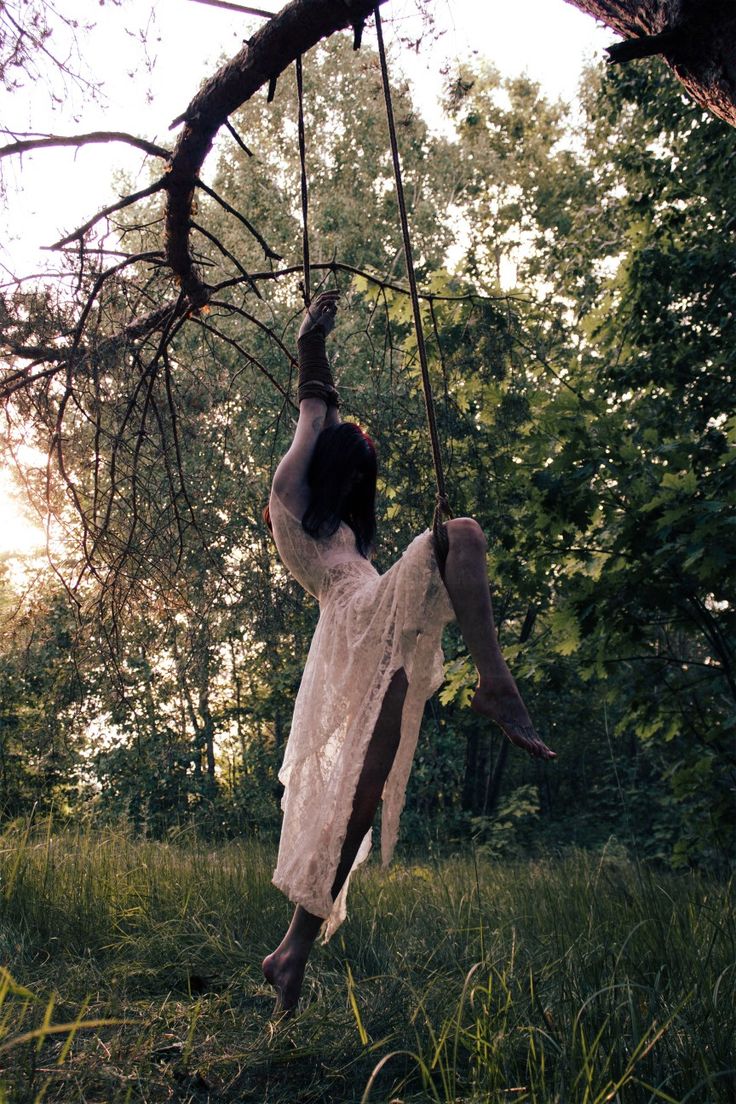 a woman in a white lace dress is suspended from a tree branch in a forest. her wrists and ankles are bound with ropes.  the scene is dimly lit, creating a mysterious and slightly unsettling atmosphere. the image has a dark, moody, and artistic feel, suggesting a theme of vulnerability or perhaps even ritualistic practice. the background is a dense forest setting, with tall trees and tall grass.