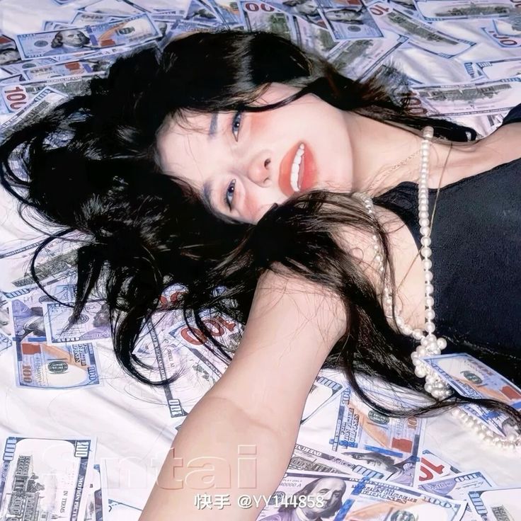 a young asian woman with long black hair and makeup lies on a bed covered in 100 dollar bills. she wears a black top and a pearl necklace. the image has a glamorous and luxurious feel.