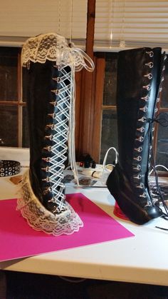 the image shows a pair of black leather platform boots with a gothic aesthetic. the boots feature intricate laceup detailing that extends up the leg, and are adorned with white lace trim. they rest on a pink surface, likely during the process of being made or modified.