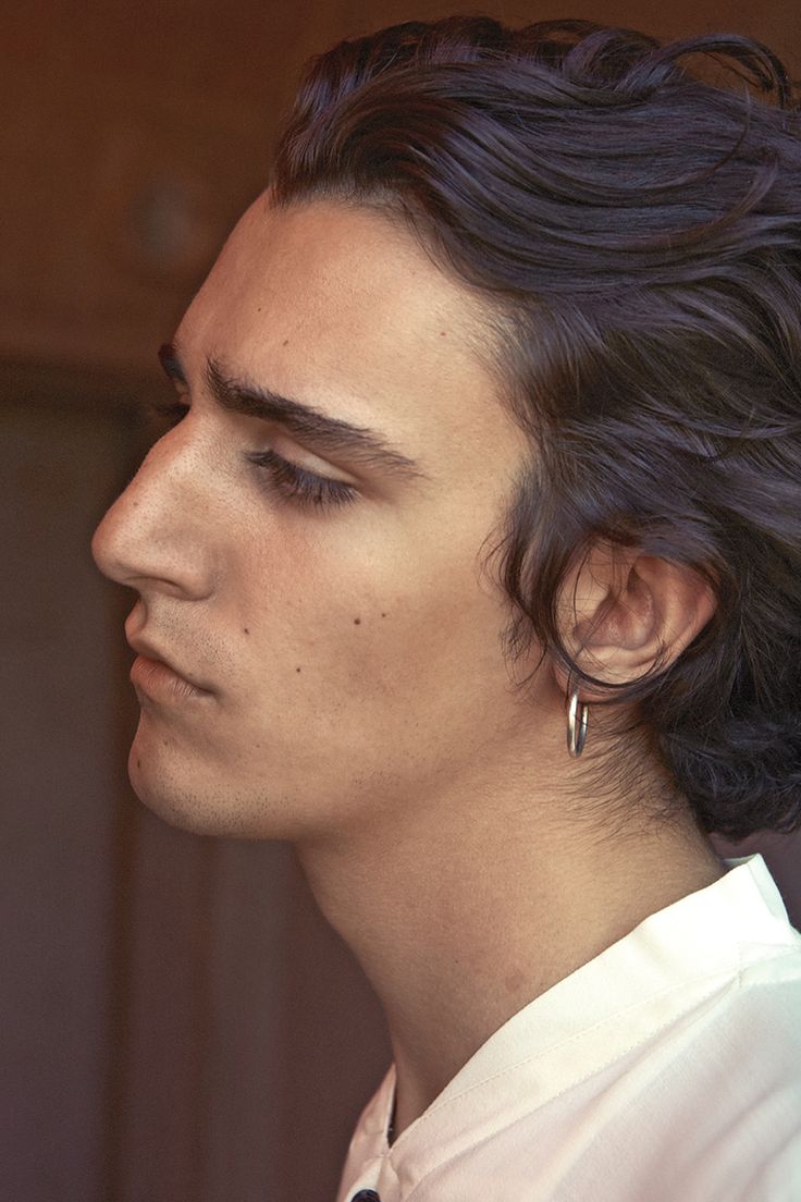 closeup profile portrait of a young man with dark, wavy hair. he is wearing a simple white shirt and a small silver hoop earring in his left ear. his expression is serious and pensive. the background is blurred and out of focus. the image is welllit and has a slightly soft focus. the overall style is fashionforward and elegant.
