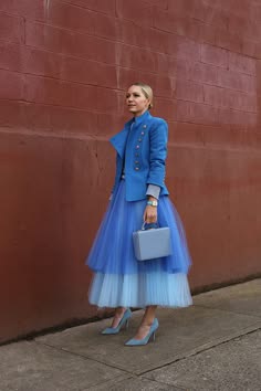 a woman wearing a blue jacket and a tulle skirt is standing in front of a red brick wall. she is carrying a blue handbag and has her hair pulled back. her heels are the same color as her skirt and handbag. she is looking to the right of the image and seems to be posing for a photo.