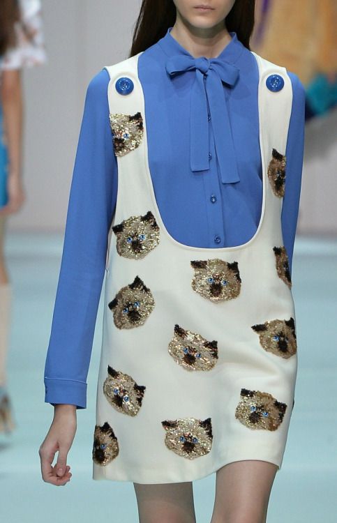 a model is wearing a light blue longsleeved blouse with a pussy bow under a sleeveless white dress featuring numerous cat face designs made with shiny fabric or sequins. the outfit is being showcased on a runway.