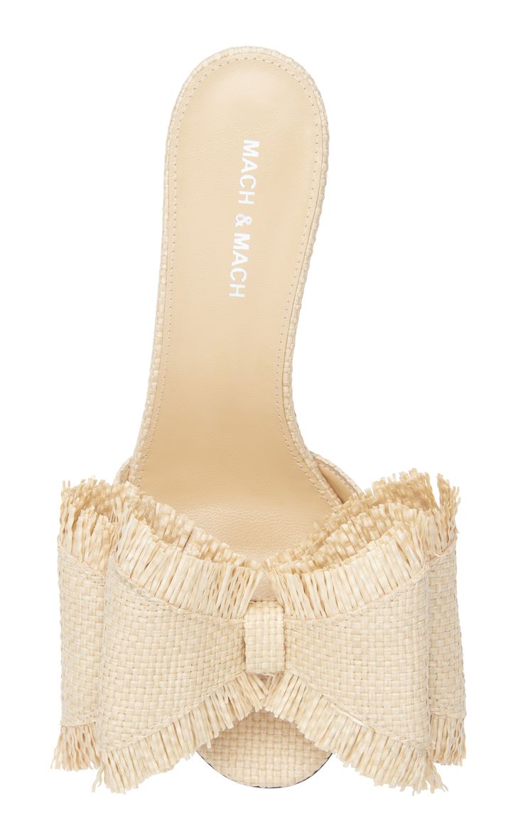 a closeup studio shot of a pair of mach & mach beige straw heeled sandals. the sandals have a large raffia bow with fringe detailing on the front.  the heel is visible and the insole shows the mach & mach branding. the background is pure white.