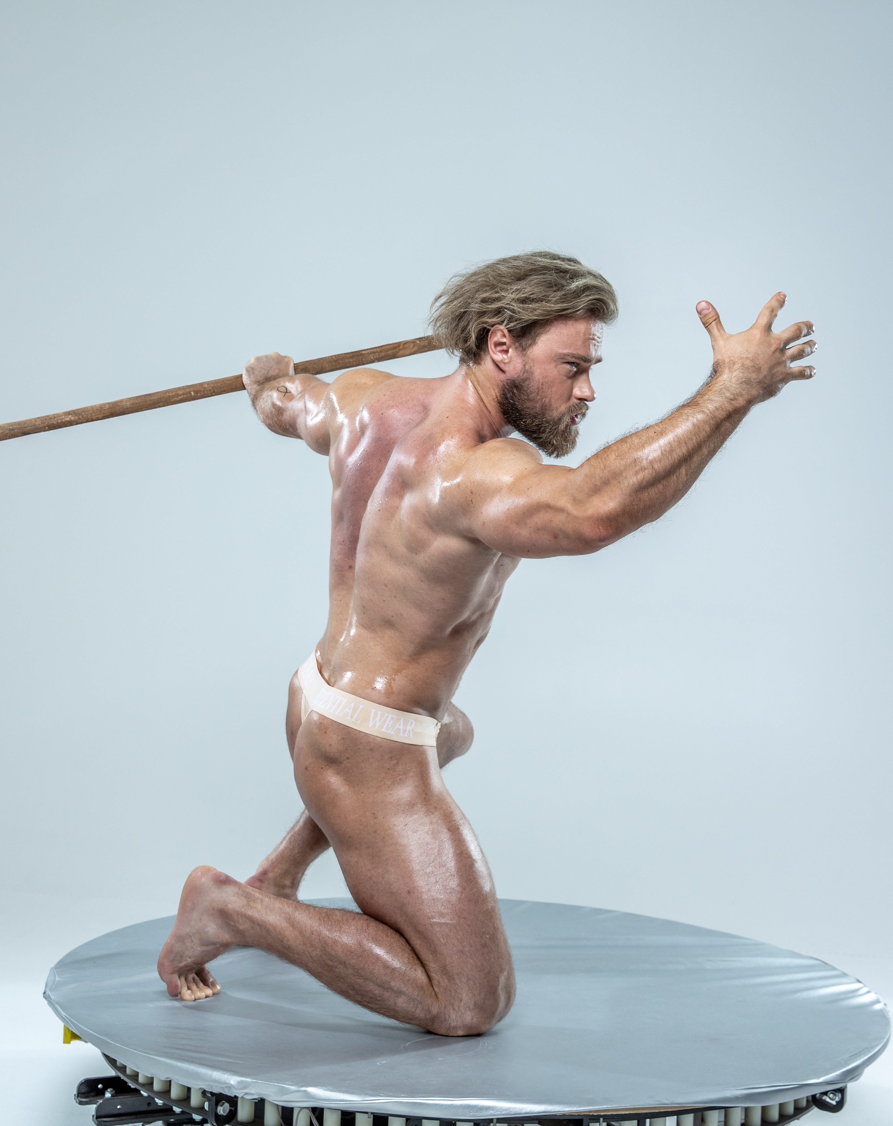 the image shows a muscular man in a dynamic pose, holding a wooden spear. he is set against a simple gray background, illuminated by soft studio lighting. his pose suggests action and power.