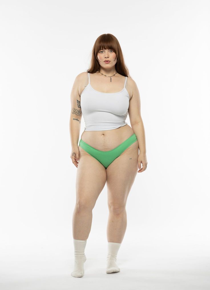a fullbody shot of a confident plussize woman with shoulderlength brown hair wearing a white tank top and light green bikinistyle briefs. she is standing with her feet slightly apart against a white background. she is wearing white socks. her expression is serious but not unfriendly. the overall image emphasizes body positivity and realistic body representation.