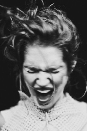 a black and white closeup portrait of a young woman with long messy hair, her mouth open wide in a yell or scream. she appears angry and furious.  the image is taken from a low angle and is in focus, with the woman's face as the focal point.  her face and expression are the main focus of the image.
