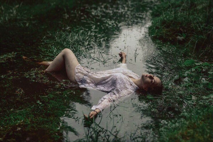 a woman is lying in a shallow stream of water, the forest surrounding her. she is wearing a white dress and her hair is wet. the water is green and the forest floor is covered in green foliage. the woman appears to be sleeping or in a state of peaceful tranquility.