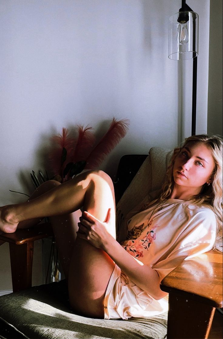 a young, fairskinned blonde woman with long hair is lounging comfortably in a wooden armchair.  she's wearing a light beige, possibly silk, pajama set with a floral print. sunlight streams into the room, illuminating her and casting a warm glow.  a vintage floor lamp with a glass shade stands behind her.  the overall atmosphere is relaxed and peaceful, with hints of stylish interior design visible.