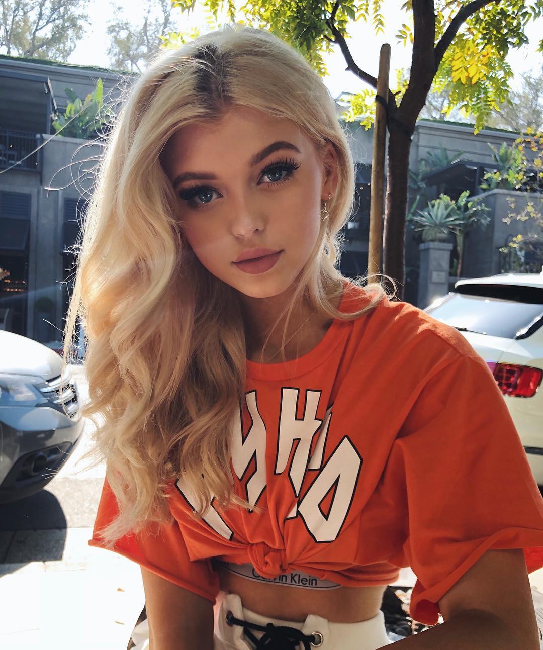 a portrait of loren gray, a young woman with blonde hair and blue eyes, wearing an orange tshirt tied at the waist, revealing calvin klein underwear. she is outdoors with cars and trees in the background.