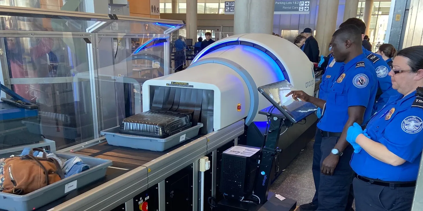 a tsa employee in a blue uniform is checking baggage with an xray scanner at an airport security checkpoint. the scanner has a conveyor belt and the employee is watching the monitor for any suspicious items. the employee is wearing a blue uniform with the tsa logo. another tsa employee is watching the screen with the first employee.