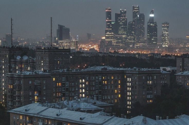 he image shows a panoramic view of moscow, russia at dusk. in the background, the modern skyscrapers of the moscow city business district are illuminated, creating a stark contrast against the darker, older residential buildings in the foreground.  the overall mood is dark and atmospheric, with a hazy quality to the air. the lights from the buildings and the city below create a sense of depth and scale. the image is taken from a vantage point that provides a sweeping view of both modern and older architectural styles, highlighting the blend of old and new in moscow's cityscape.