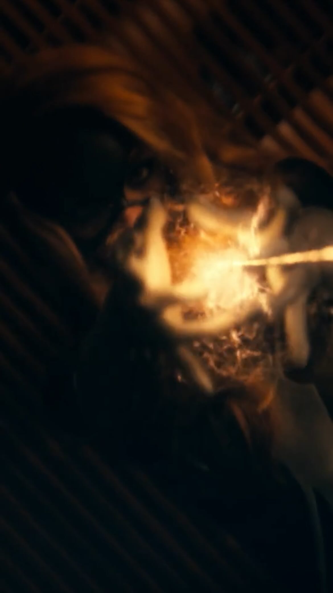 the image is a dark, blurry closeup shot of what appears to be blonde hair on fire. the flames are intense and bright against the dark background. the overall impression is one of chaos and destruction, possibly suggesting an accident or a dramatic scene. the image is grainy and has a filmlike quality, enhancing its gritty and intense atmosphere. the focus is soft, making some details unclear, but the burning hair is the central focus.