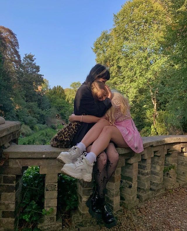 two girls are sitting on a stone wall outdoors. the girl on the left is wearing white sneakers and the girl on the right is wearing fishnet tights and a pink skirt. they are hugging each other and smiling. the girls are surrounded by green trees and a blue sky. they seem to be enjoying their time together.