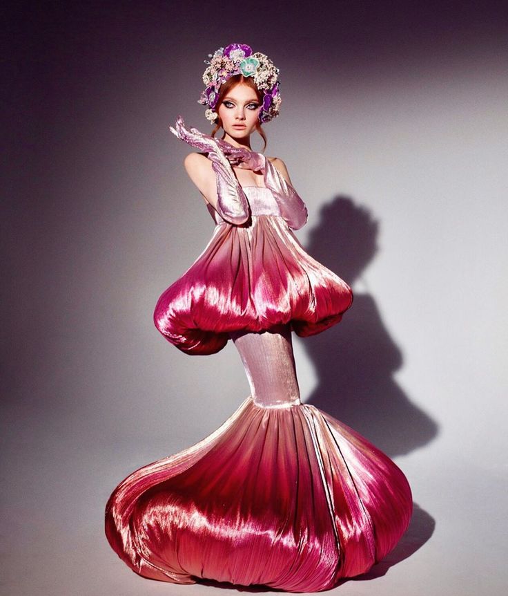 a model with red hair and red lips is wearing a beautiful  pink dress.  the  dress is  a  silky,  fluid  fabric that  appears to be  shaped  like  a  flower.  the  model  has a flower crown in her hair.  she is  posing  in a studio with a soft,  purple  background.  she  is  looking  at  the  camera.  she  is  wearing  long,  pink  gloves.  the  model  looks  elegant  and  glamorous.  her makeup is bold and eyecatching.  the overall effect of the image is one of  style and  beauty.
