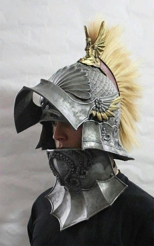 a man models a metallic roman helmet with intricate details, including a blonde mohawk crest and sculpted figures. the helmet is made of a dark, metallic material and has a weathered appearance. the man is wearing a black shirt and is standing against a white background.