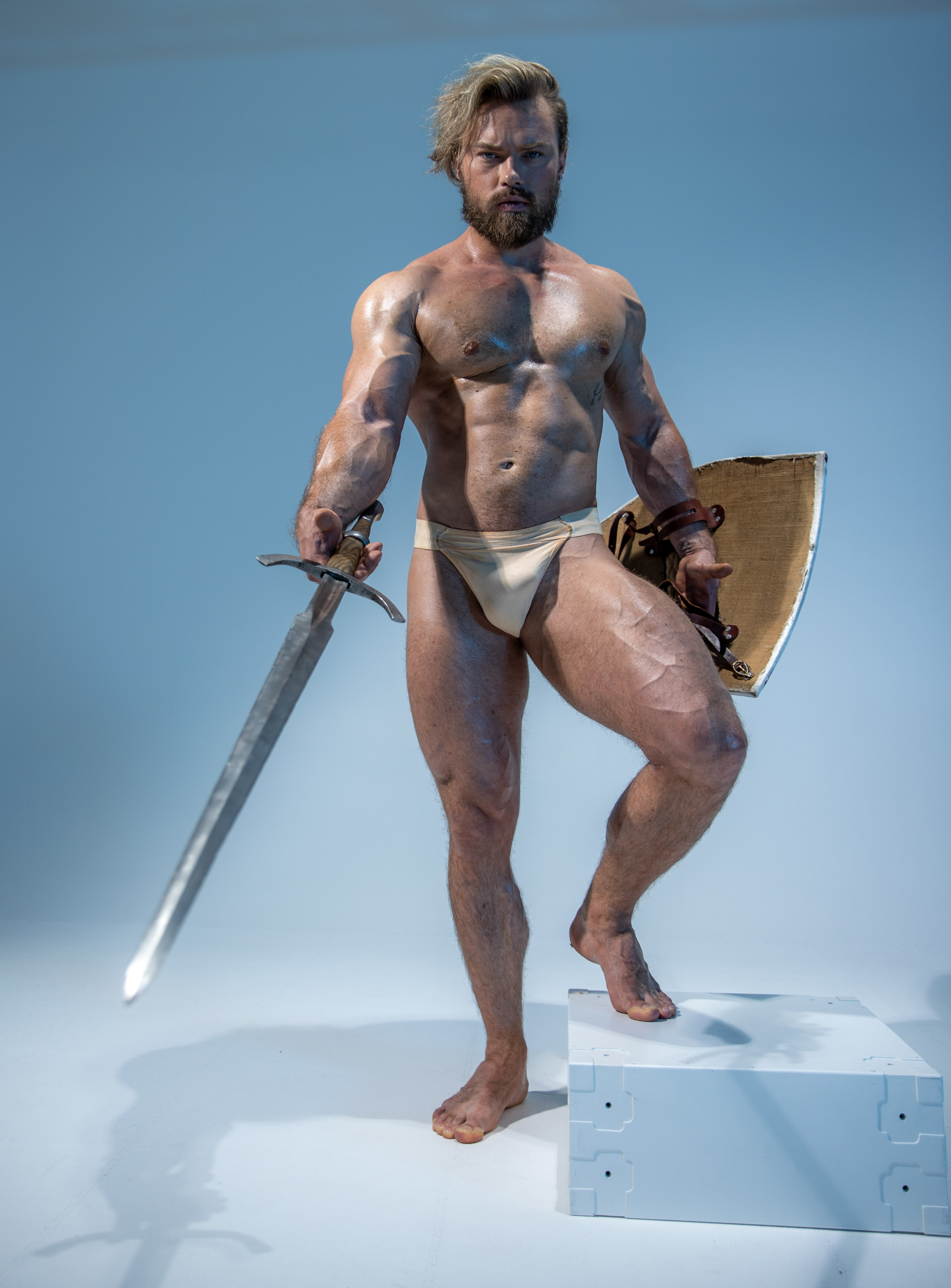 a muscular man with long blond hair and beard stands in a confident pose against a light blue studio backdrop. he wears only a light beige speedo and is covered in a light sheen of sweat, highlighting his physique. he holds a gleaming sword in his right hand, its point directed towards the ground. in his left hand, he grips a round shield with leather straps and metal accents. he stands with one foot on a white cube, further enhancing his powerful stance.