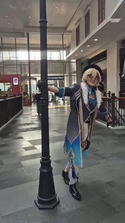 a male cosplayer dressed as a fictional character stands in a city mall by a lamppost. they are smiling and looking to the left. the cosplayer is wearing a blue, black, and white costume with feather and fur accents.