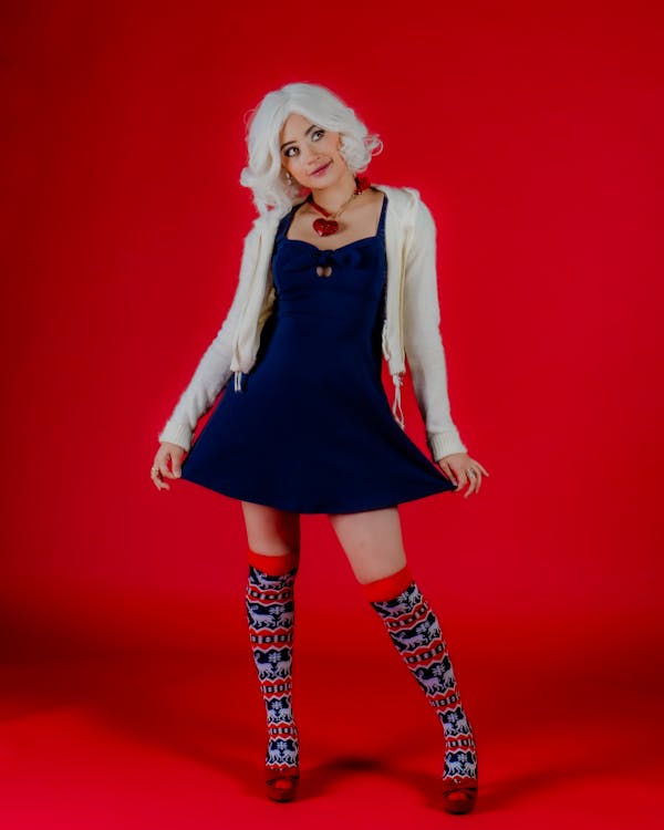 a woman stands against a bright red background. she has white, curly hair and wears a dark blue dress, a cream colored cardigan, and red shoes. she has on knee socks with a pattern, and a heart necklace. she has one hand on her dress.