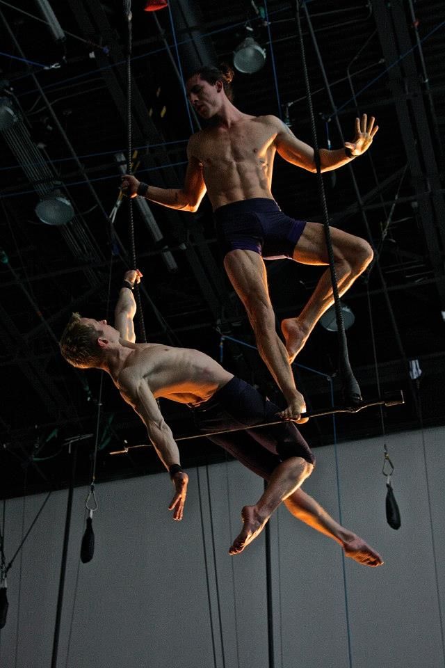 two shirtless men are performing on a trapeze in a dark gym. the man on top is holding onto the trapeze bar with his left hand while his right hand is raised in the air. the man on the bottom is hanging from his left hand on the trapeze and is in the process of swinging up. the image is lit from the top, casting shadows on the men's bodies. the background is dark with wires and ropes suspended from the ceiling.