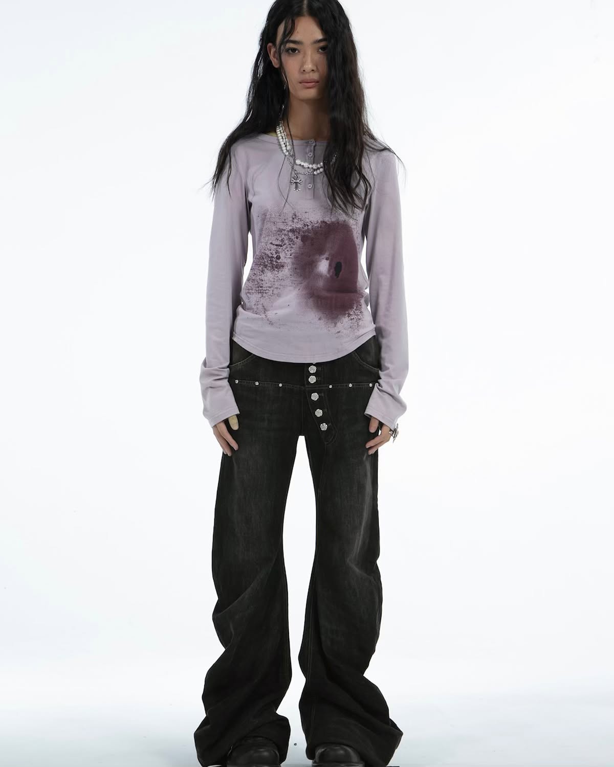 a fullbody studio shot of a female model against a white background. she is wearing a pale lavender longsleeved shirt with a dark purplishbrown paint splatter design across the chest. the shirt has a relaxed, slightly oversized fit. she's also wearing highwaisted, wideleg dark wash jeans with a button fly and subtle stud detailing along the waistline.  the jeans have a flared or bellbottom shape. she's accessorized with a simple pearl necklace. the overall style is a blend of contemporary and grunge influences. the model is standing with a neutral expression.