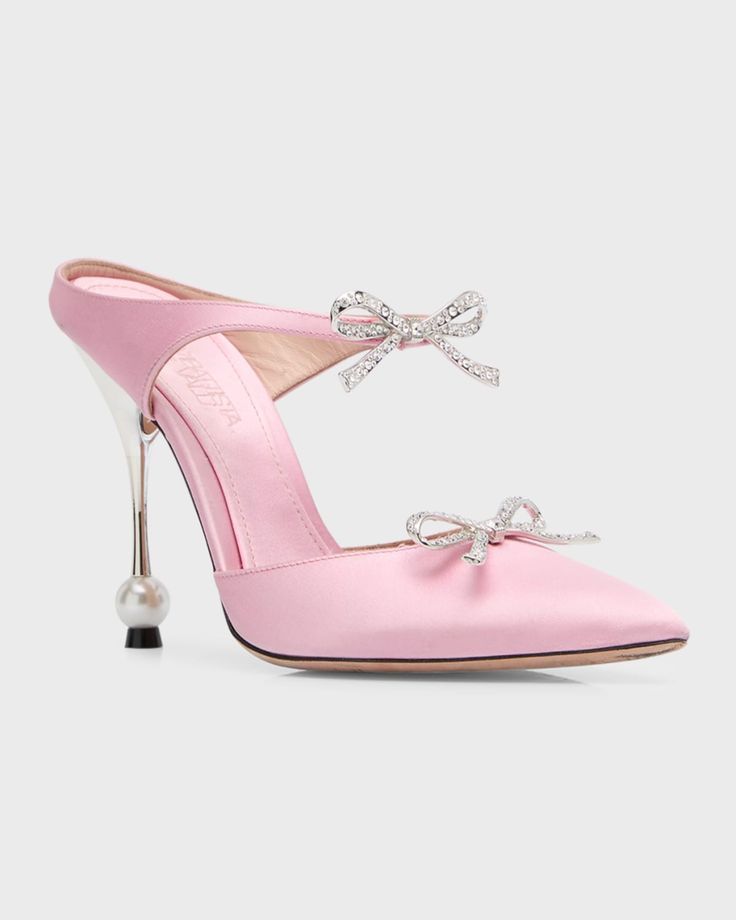 a closeup studio shot showcases a pink satin mulestyle shoe adorned with a decorative bow on the upper and a clear stiletto heel with a pearl detail at the base.