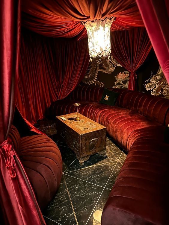 a cozy and intimate lounge with plush red velvet couches and a lowlying coffee table. the room is dimly lit with a single chandelier hanging from a red velvet canopy, creating a dramatic and luxurious atmosphere. the black and red color scheme is sophisticated and elegant, while the soft lighting creates a sense of privacy and intimacy. the couch is arranged in a curved shape that invites guests to relax and converse, while the low coffee table creates a sense of closeness and intimacy. the entire space exudes an air of luxury and sophistication, making it the perfect place to relax and unwind.