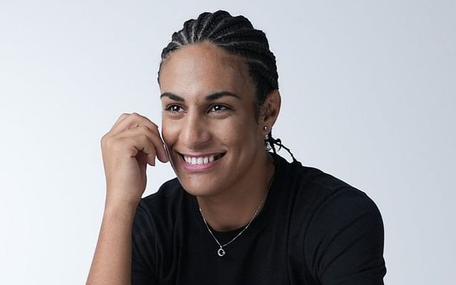 a smiling woman with braided hair is wearing a black tshirt and a silver necklace. the background is white and the lighting is soft.