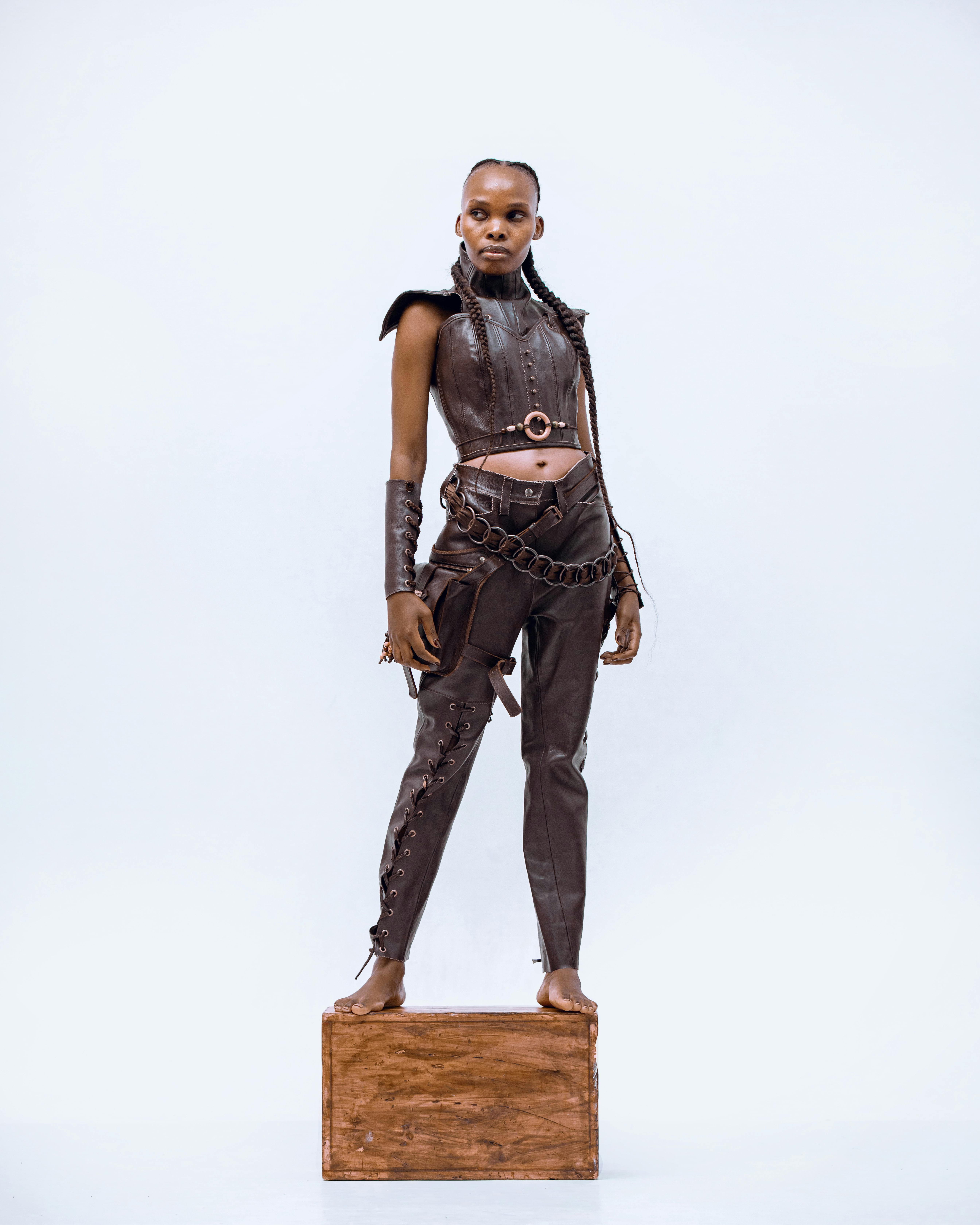 a woman stands barefoot on a wooden box, modeling a brown leather outfit consisting of a cropped top, pants, and accessories such as arm cuffs and a chain belt. she has braids in her hair and is positioned in a fullbody shot against a white background.