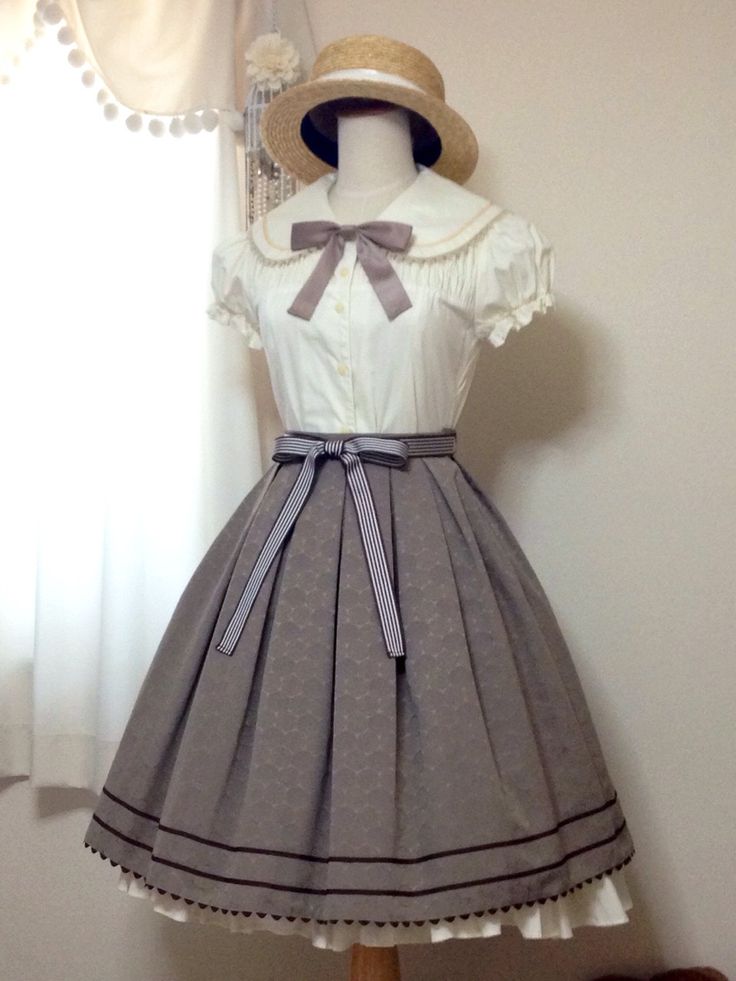 a full shot of a lolita fashion dress on a mannequin. the dress features a white blouse with a large sailor collar, brown ribbon, and puffy sleeves, paired with a highwaisted brown skirt with a subtle pattern. the skirt has pleats and a brown trim along the hemline.  a straw boater hat sits atop the mannequin's head.  the background is a creamcolored wall with a sheer white curtain.