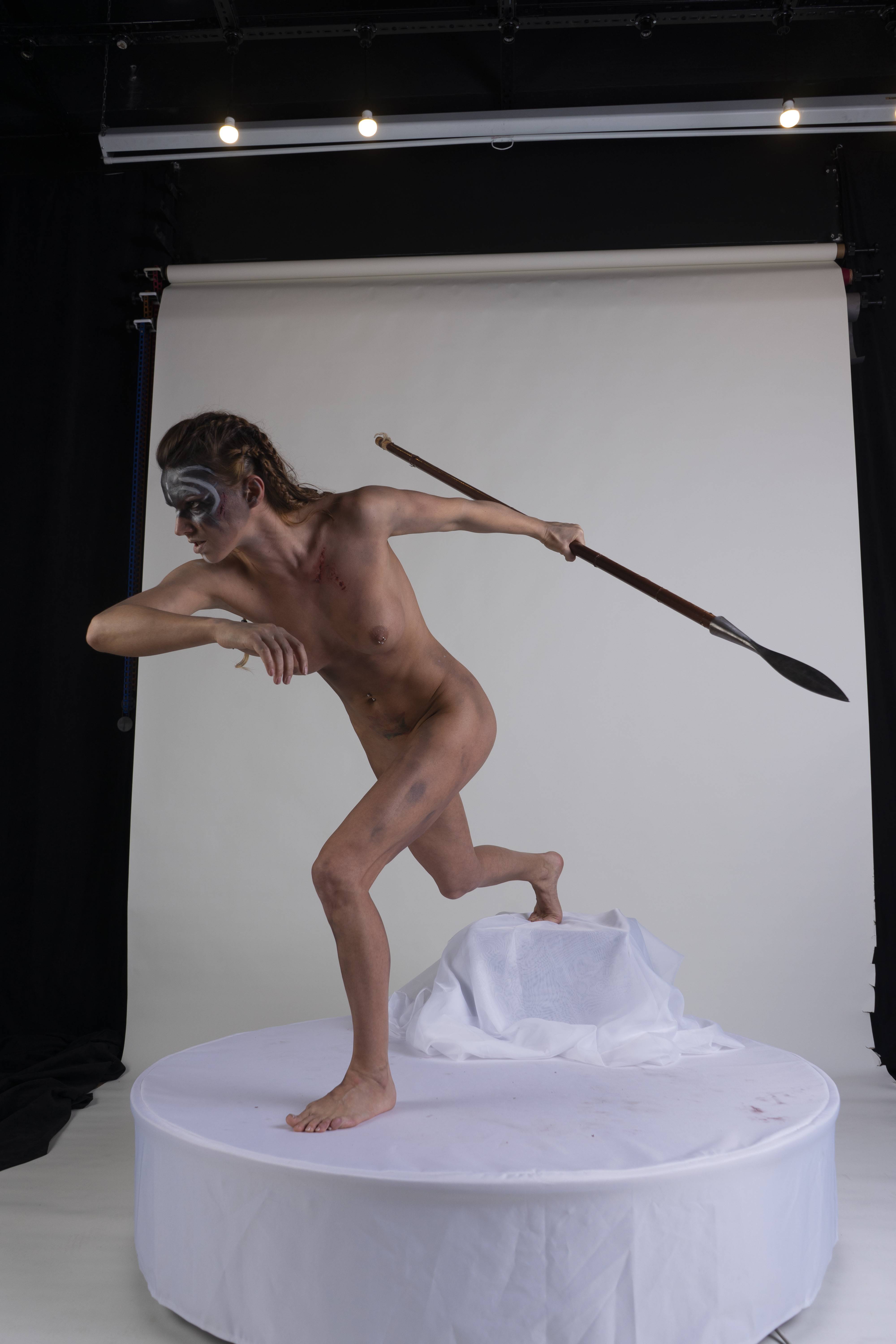 a nude woman with warrior makeup stands on a white platform, holding a spear in an action pose against a white backdrop in a studio setting.