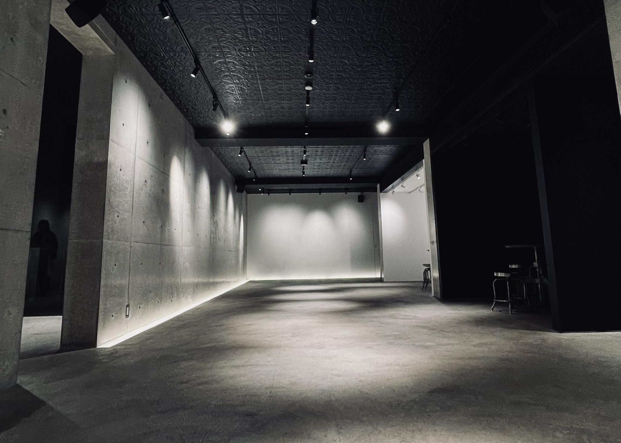 the image shows an empty studio space with a minimalist industrial design. the walls are concrete, the ceiling is black, and the floor is polished concrete. track lighting provides illumination.  the overall color scheme is grayscale, with shades of black, gray, and white dominating.