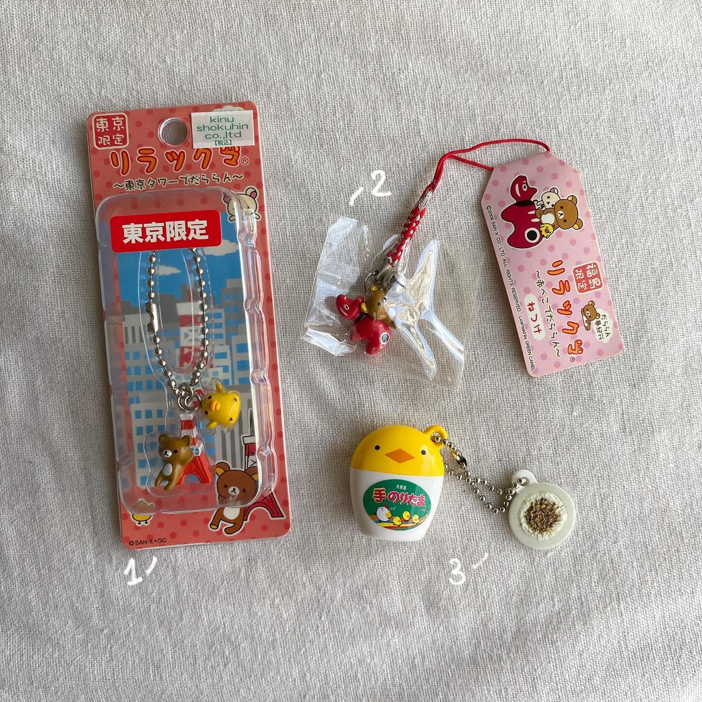 the image shows a collection of japanese character goods featuring rilakkuma, a popular bear character from sanx, and other related items. there's a packaged keychain of rilakkuma and a chick in front of a tokyo tower background, another rilakkuma keychain with red accessories still in its plastic packaging, and a yellow chickshaped keychain attached to a replica of a condiment container with the words "手のりたま" (tenori tama) written on it. a rilakkuma themed tag accompanies one of the keychains.