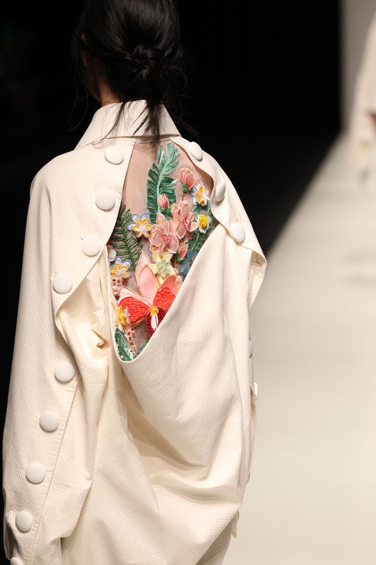 the image shows a closeup view of a model's back, showcasing a unique and intricately designed garment. the outfit is a creamcolored shirt with large buttons running down the sleeves and back. the back of the shirt features a sheer panel adorned with a striking, embroidered floral arrangement. the floral embroidery is highly detailed and uses a variety of colors and textures. the overall effect is a blend of sophisticated simplicity with a striking artistic element. the model's hair is pulled back neatly, allowing for a full view of the intricate back detail.  the runway background is visible, indicating this is likely a fashion show photograph.