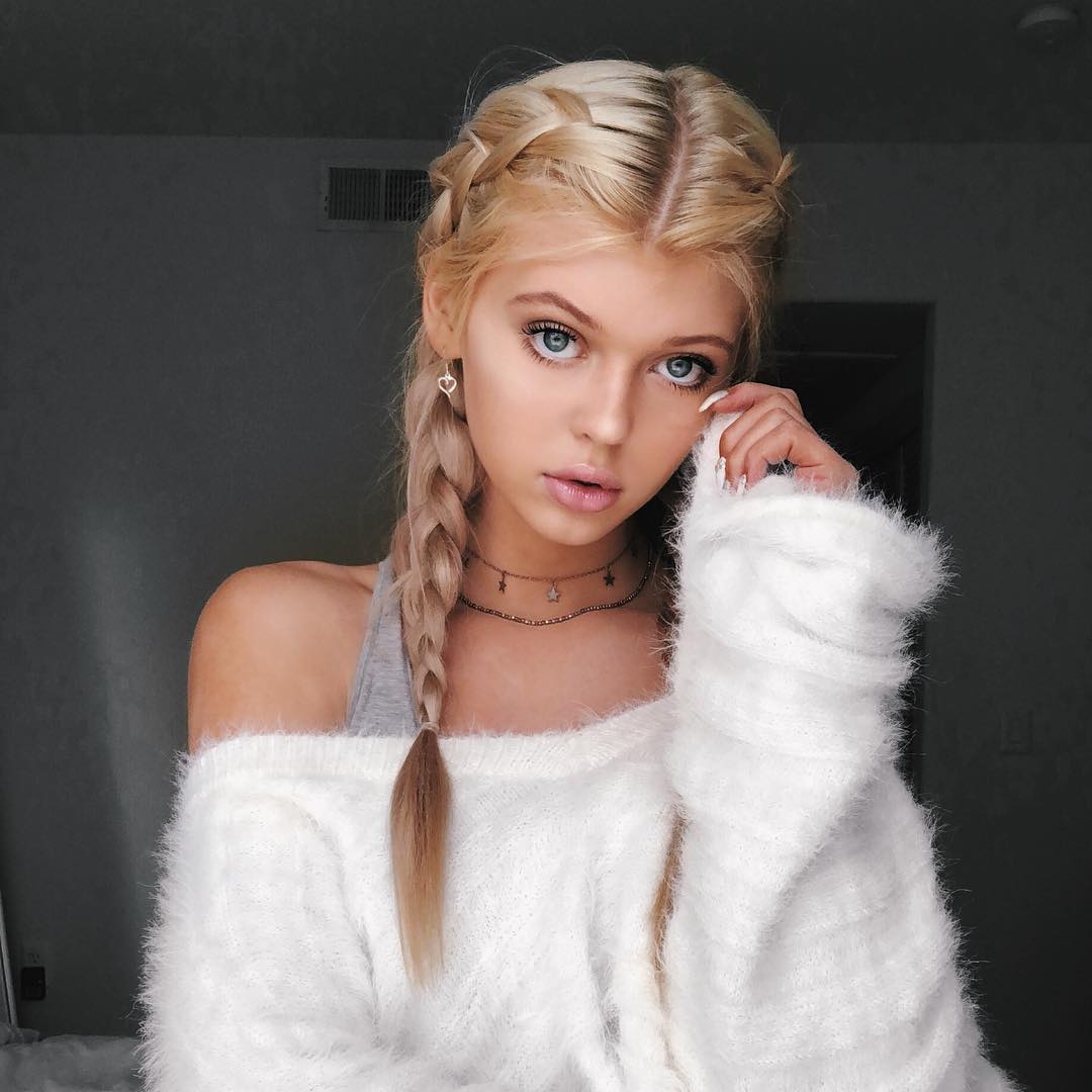 the image shows a young woman with blonde hair styled in braids. she is wearing a white, fuzzy sweater and a delicate necklace. she has striking blue eyes. the background is a blurred, neutral color. she is wearing heart shaped earrings.