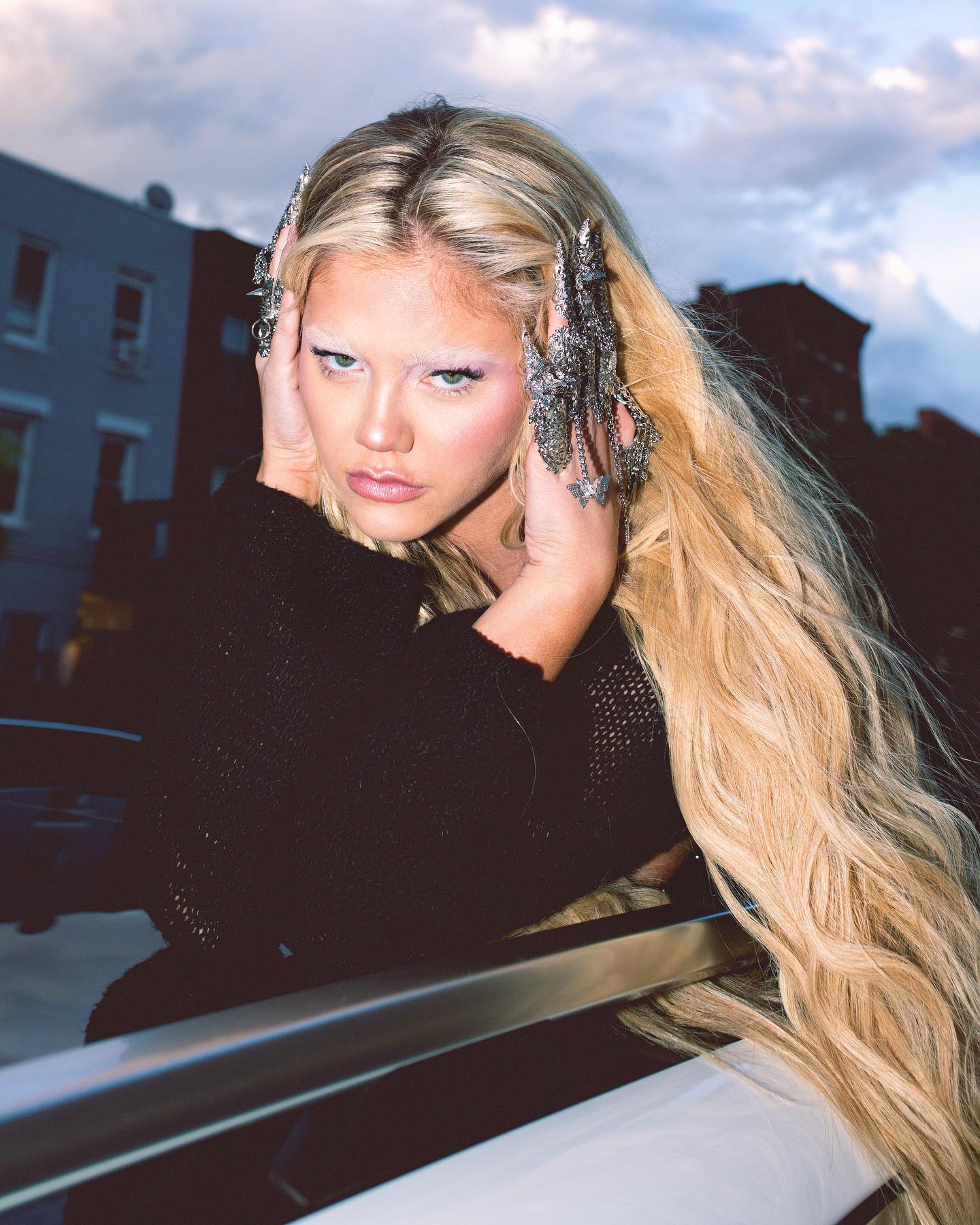 a woman with long blonde hair, pale skin and light eyebrows is shown in an outdoor setting. she is wearing black sweater. the woman is leaning on a metallic bar, with buildings and an evening sky in the background. she is wearing elaborate jewelry on her fingers. the makeup on her face is noticeable.