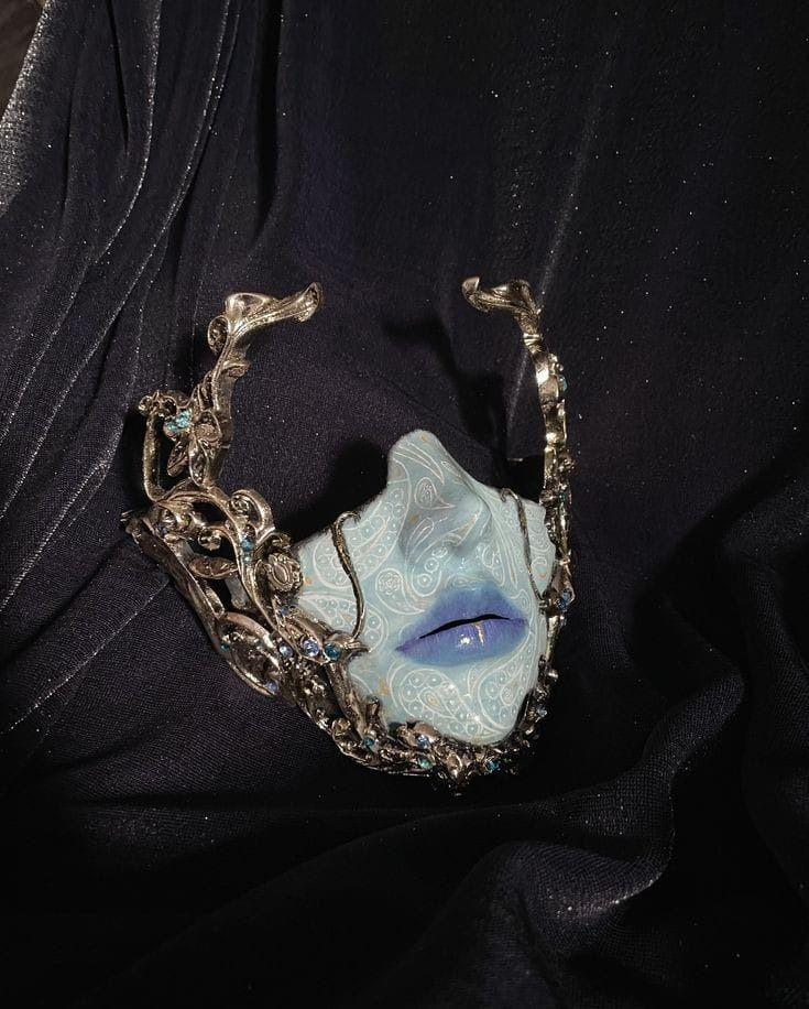 a handcrafted silver mask lays on dark blue velvet fabric. the mask is crafted to look like a face with blue cracked skin and blue lips. small blue gems are embedded in the ornate silver.